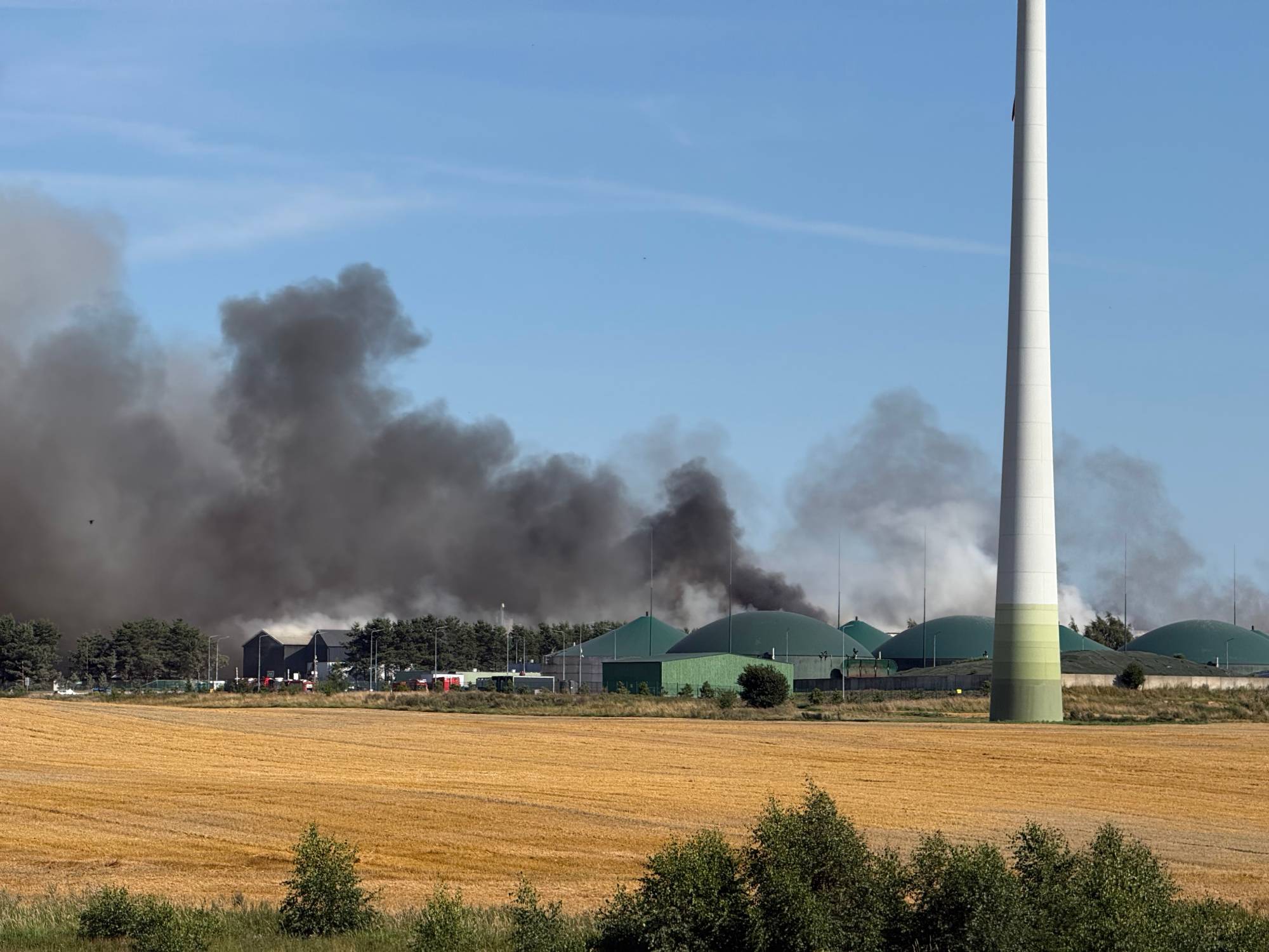 Widok z oddali na pożar zakładu przetwarzania odpadów w Chlewnicy. Na pierwszym planie rozciąga się skoszone, żółte pole oraz podstawa wielkiej turbiny wiatrowej. W tle, za zielonymi kopułami biogazowni i halami, unoszą się gęste, ciemne kłęby dymu na tle błękitnego nieba