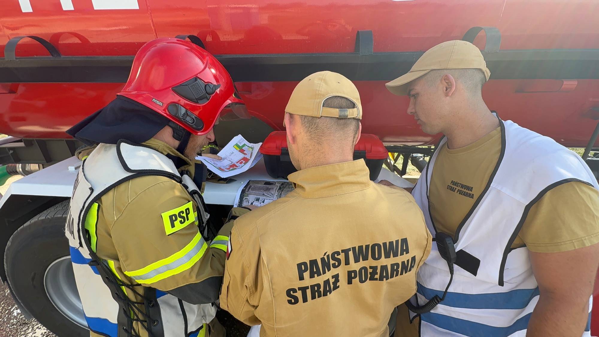 Trzej strażacy Państwowej Straży Pożarnej w piaskowych mundurach stojący przy czerwonym wozie gaśniczym i analizujący mapy oraz plany techniczne obiektów. Jeden ze strażaków ma na głowie czerwony hełm bojowy, dwaj pozostali beżowe czapki z daszkiem. Kilkadziesiąt jednostek straży pożarnej prowadzi skomplikowaną akcję obrony zagrożonych hal i tłumienia ognia na terenie zakładu ELWOZ w Chlewnicy
