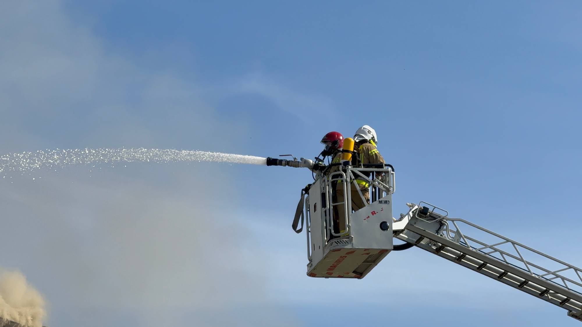 Strażacy gaszą pożar w Chlewnicy z wykorzystaniem podnośnika hydraulicznego. Dwaj strażacy w pełnym ekwipunku bojowym i aparatach oddechowych stoją w koszu podnośnika na wysuniętej drabinie pożarniczej na tle nieba. Z zamontowanego działka kierują silny, rozproszony strumień wody gaśniczej w stronę zarzewia ognia.