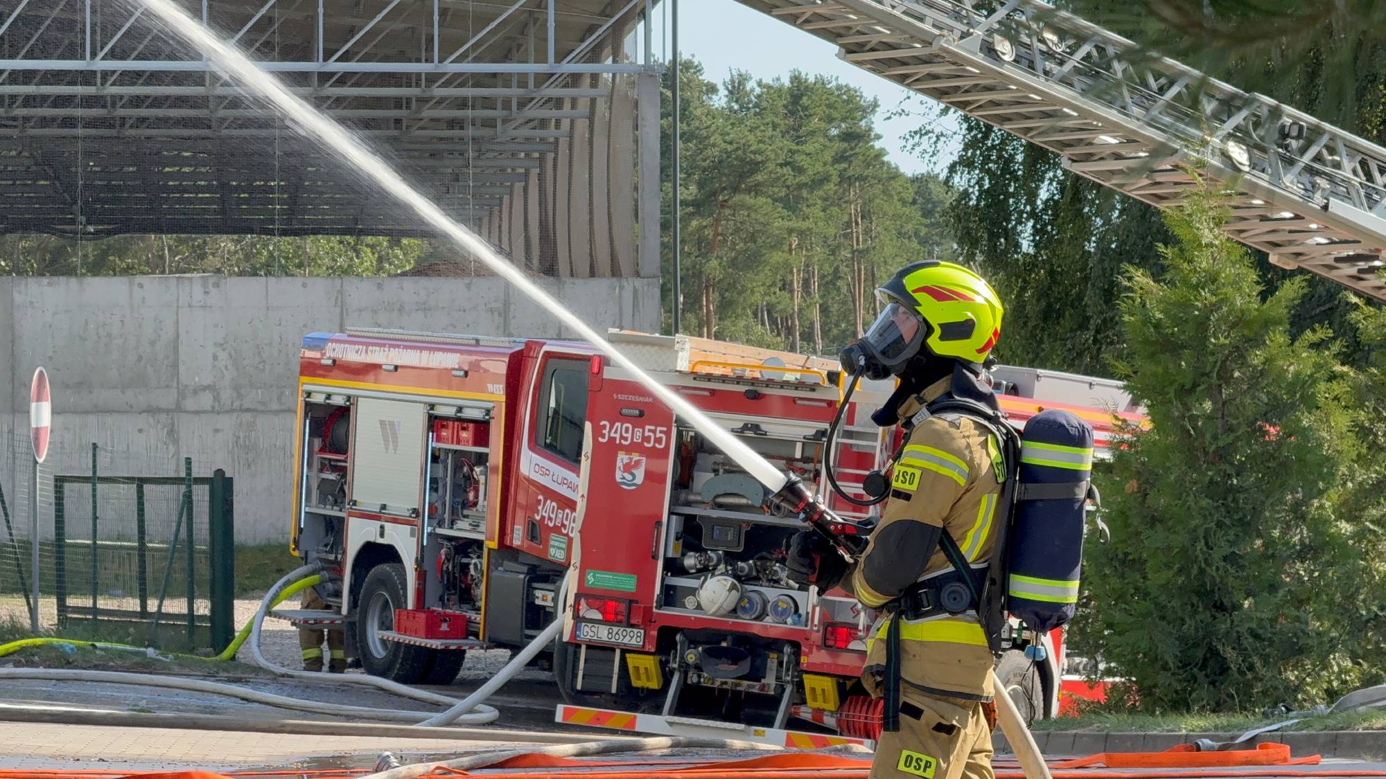 Strażak w piaskowym kombinezonie bojowym, masce oddechowej i jaskrawożółtym hełmie kieruje silny strumień wody z prądownicy ku górze. W tle stoi czerwony wóz strażacki OSP Łupawa z otwartymi skrytkami sprzętowymi, a nad nim widoczna jest wysunięta drabina mechaniczna. Strażacy z lokalnych jednostek OSP i PSP biorą udział w akcji gaśniczej w Chlewnicy w powiecie słupskim