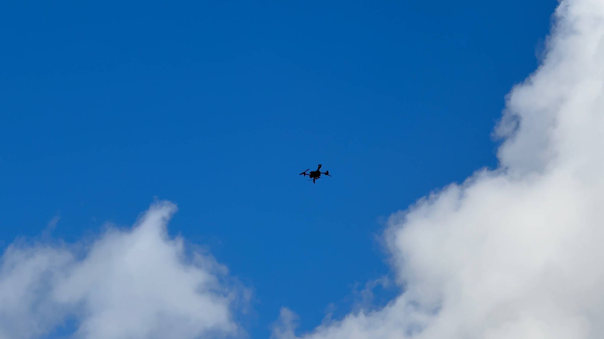 Widok z dołu na ciemną sylwetkę drona lecącego na tle błękitnego nieba z białymi, pierzastymi chmurami. Dron z kamerą termowizyjną sprawdza, czy pożar na terenie zakładu przetwarzania odpadów w Chlewnicy został już całkowicie dogaszony