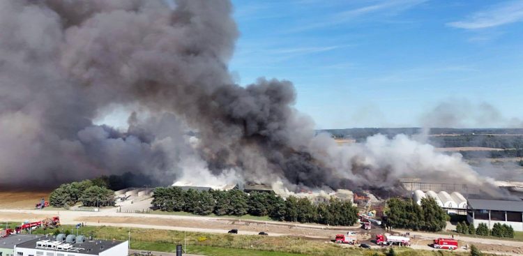 Zdjęcie z drona przedstawiające pożar zakładu przetwarzania odpadów w Chlewnicy. Nad rzędem hal unoszą się olbrzymie, kłębiące się masy czarnego i białego dymu zasłaniające część nieba. Na drogach dojazdowych i wokół terenu zakładu widać rozstawione liczne czerwone wozy straży pożarnej
