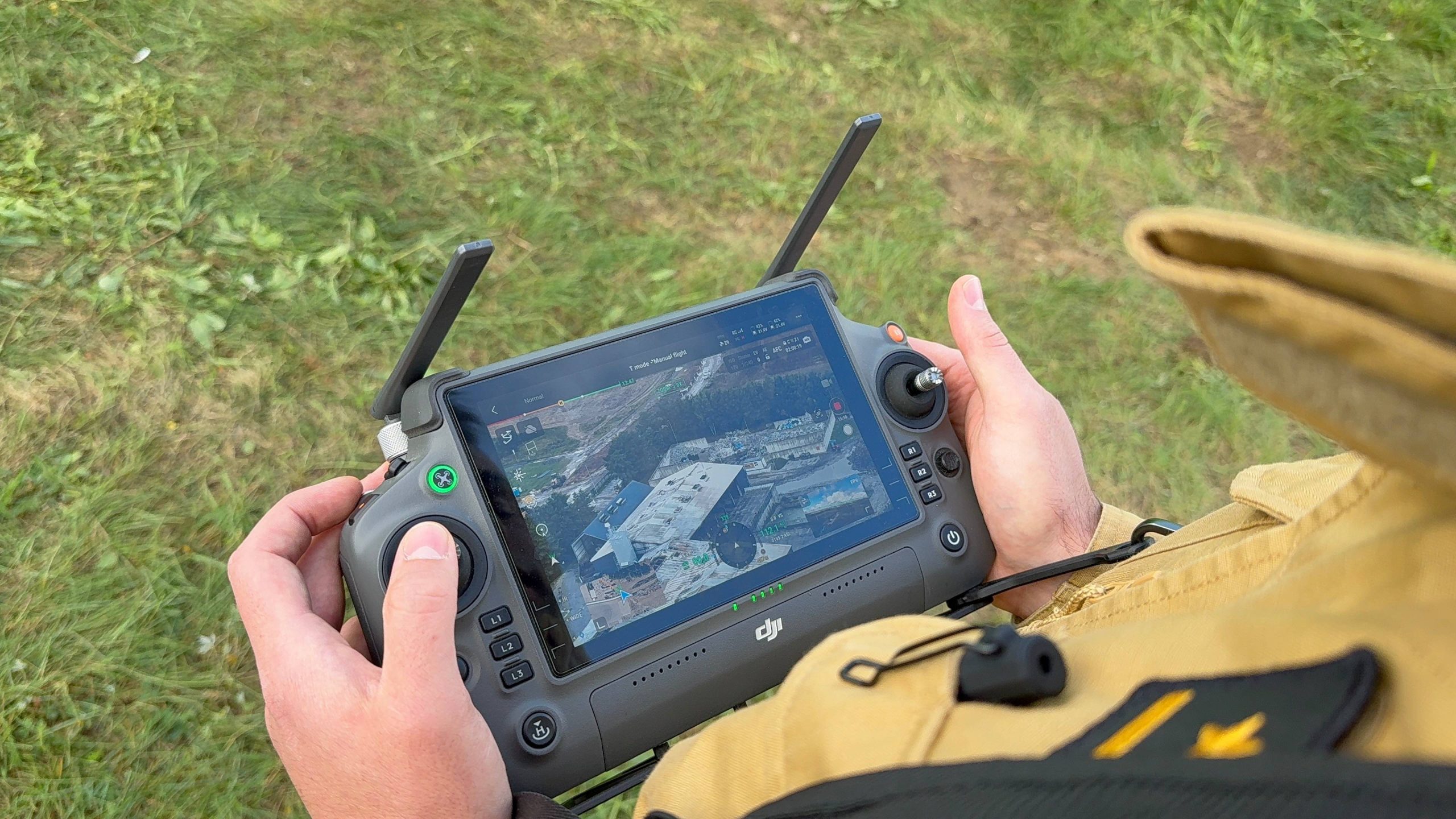 Widok z góry na ręce osoby w piaskowym mundurze strażackim, która trzyma aparaturę sterującą dronem (aparatura DJI z wbudowanym ekranem). Na wyświetlaczu widoczny jest podgląd z kamery drona przedstawiający zniszczone przez pożar hale i pogorzelisko zakładu w Chlewnicy