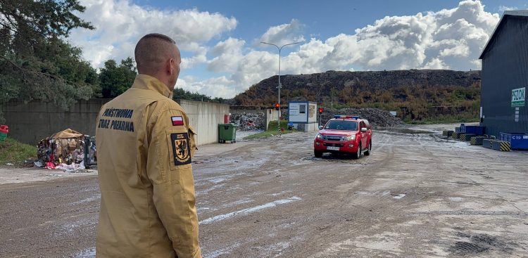 Strażak w jasnobrązowym mundurze z napisem „Państwowa Straż Pożarna” na plecach i naszywką „KM PSP Słupsk” stoi tyłem. Patrzy na teren zakładu przetwarzania odpadów w Chlewnicy, po którym porusza się czerwony samochód ratowniczy. W tle widoczna jest hałda odpadów i budynek PSZOK