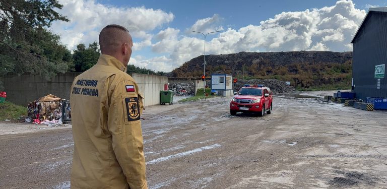 Strażak w jasnobrązowym mundurze z napisem „Państwowa Straż Pożarna” na plecach i naszywką „KM PSP Słupsk” stoi tyłem. Patrzy na teren zakładu przetwarzania odpadów w Chlewnicy, po którym porusza się czerwony samochód ratowniczy. W tle widoczna jest hałda odpadów i budynek PSZOK