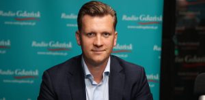 Przemysław Majewski w studiu Radia Gdańsk. Gdański radny w ciemnej marynarce i jasnej koszuli w paski patrzy w stronę obiektywu, siedząc na tle turkusowej ścianki z powtarzającym się logo Radia Gdańsk