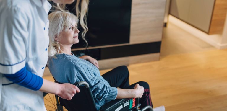 Starsza kobieta w niebieskim swetrze siedząca na wózku inwalidzkim, obok której stoi opiekunka w białym fartuchu medycznym. Zapowiedź powstania budynku opieki wytchnieniowej w Pucku