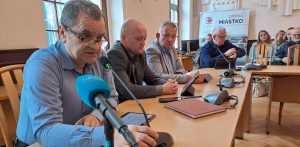Kilku mężczyzn siedzi przy drewnianym stole w sali konferencyjnej podczas spotkania dotyczącego kryzysu w szpitalu w Miastku. Na pierwszym planie w niebieskiej koszuli i okularach pochyla się nad mikrofonem radny Dariusz Zagaja, obok niego siedzą inni uczestnicy spotkania, a w tle widać transparent z napisem "Gmina Miastko". Radny chce odwołania burmistrza