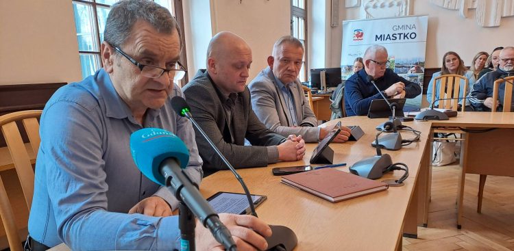 Kilku mężczyzn siedzi przy drewnianym stole w sali konferencyjnej podczas spotkania dotyczącego kryzysu w szpitalu w Miastku. Na pierwszym planie w niebieskiej koszuli i okularach pochyla się nad mikrofonem radny Dariusz Zagaja, obok niego siedzą inni uczestnicy spotkania, a w tle widać transparent z napisem "Gmina Miastko". Radny chce odwołania burmistrza