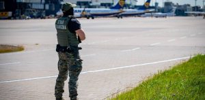 Tyłem do obiektywu stoi funkcjonariusz Morskiego Oddziału Straży Granicznej w zielonym mundurze i kamizelce taktycznej z napisem "Straż Graniczna" na plecach. W tle widoczna jest płyta lotniska z zaparkowanymi samolotami