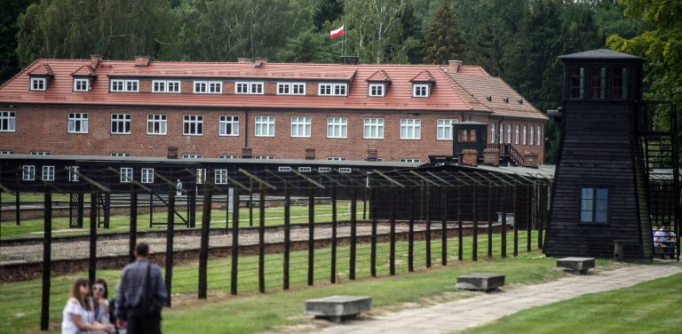 Teren byłego niemieckiego nazistowskiego obozu koncentracyjnego Stutthof w Sztutowie. Na pierwszym planie widoczne są ogrodzenie z drutu kolczastego na słupkach, drewniana wieża strażnicza oraz zwiedzający obóz ludzie. W tle znajduje się budynek z czerwoną dachówką i polską flagą na dachu. Ministerstwo Kultury i Dziedzictwa Narodowego ogłosiło konkurs na stanowisko dyrektora Muzeum Stutthof w Sztutowie