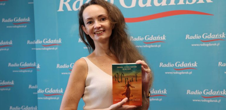 Kobieta z brązowymi, kręconymi włosami, ubrana w jasny top, stoi na tle niebieskiej ścianki reklamowej Radia Gdańsk. Uśmiecha się do aparatu i trzyma w rękach książkę zatytułowaną "Upał". To tancerka orientalna i pisarka Marta Nowicka-Gawior