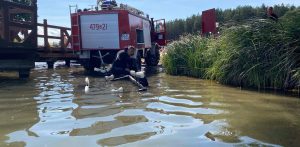 Czerwony wóz strażacki zaparkowany na brzegu jeziora w Siemirowicach tuż obok drewnianego pomostu i szuwarów. W wodzie stoi nurek w piance neoprenowej rozmawiający przez telefon, opierając się o częściowo zanurzoną przyczepę podłodziową. Z jeziora wydobyto ciała kobiety oraz mężczyzny. Kobieta była w ciąży