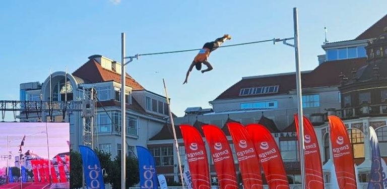 Skok o tyczce podczas mityngu "Tyczka na molo" 2026 w Sopocie - zawodnik w locie pokonuje poprzeczkę na tle sopochich budynków i czerwonych flag sponsorów