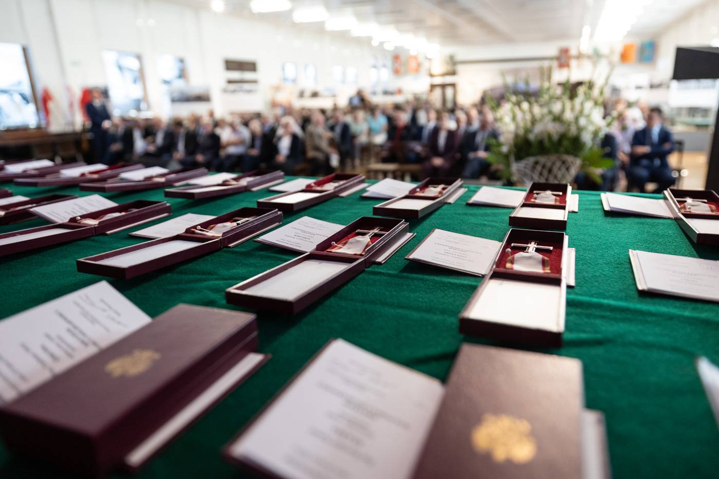 Widok z bliska na stół pokryty zielonym suknem, na którym leżą otwarte bordowe etui z Krzyżami Wolności i Solidarności oraz dokumenty potwierdzające ich nadanie. W tle, w głębi jasnej sali, widoczni są siedzący uczestnicy uroczystości