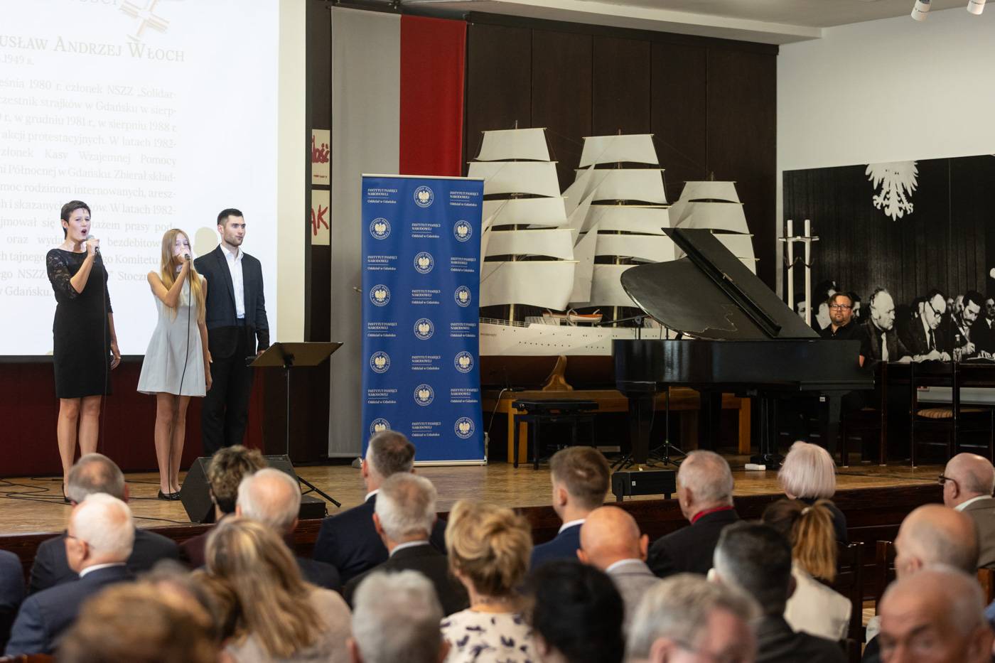 Uroczystość wręczenia Krzyży Wolności i Solidarności w Sali BHP w Gdańsku. Trójka młodych artystów – dwie kobiety i mężczyzna – śpiewa na scenie w Sali BHP w Gdańsku podczas występu artystycznego. Obok nich przy fortepianie siedzi akompaniator. Na widowni siedzą uczestnicy uroczystości, patrząc w stronę sceny. W tle widoczny jest duży model żaglowca oraz ścianki z logotypami IPN