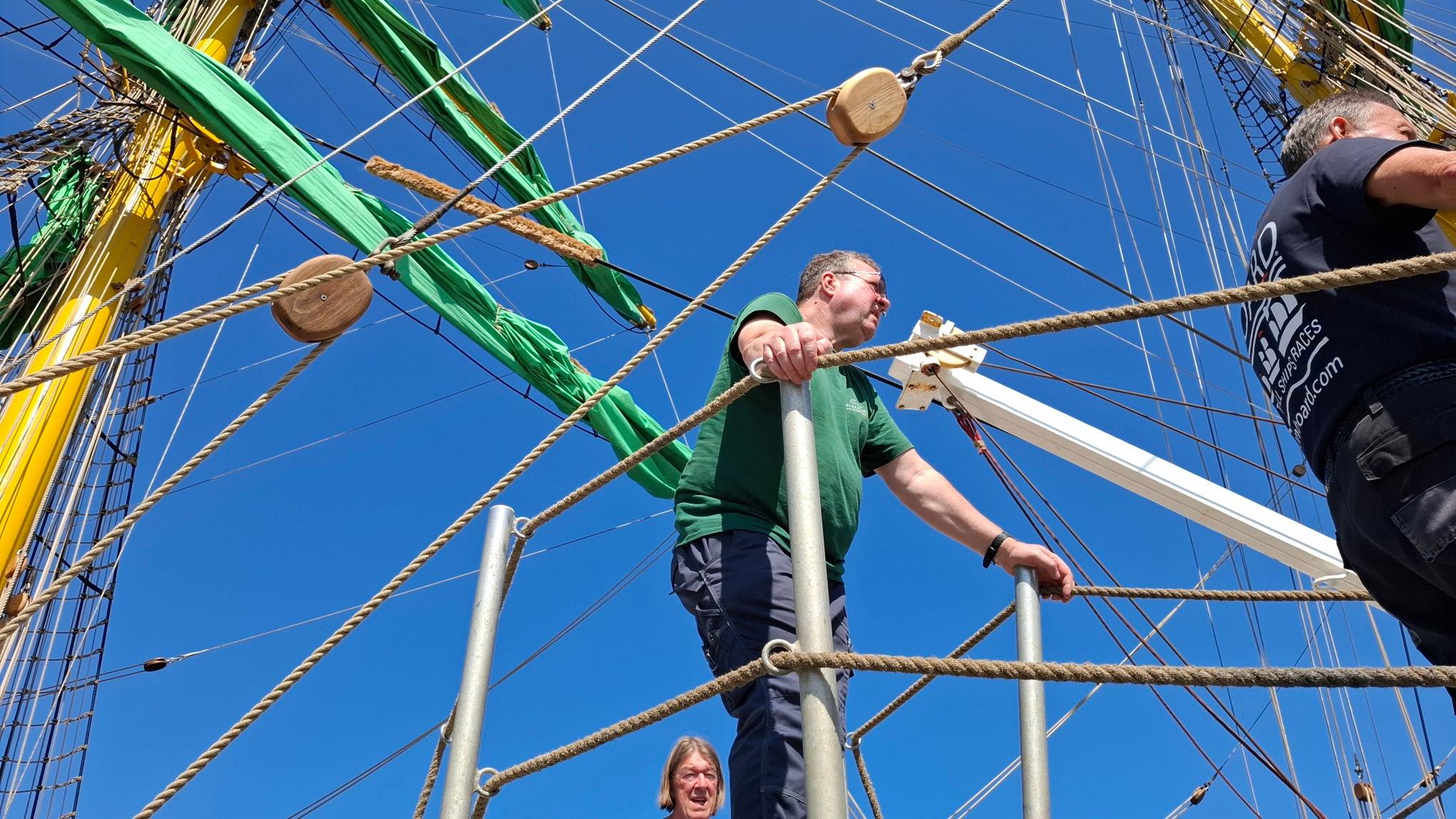 Członkowie załogi na pokładzie żaglowca Alexander von Humboldt II. W tle widoczne są liny, takelunek, żółte maszty, zwinięte zielone żagle oraz bezchmurne, niebieskie niebo
