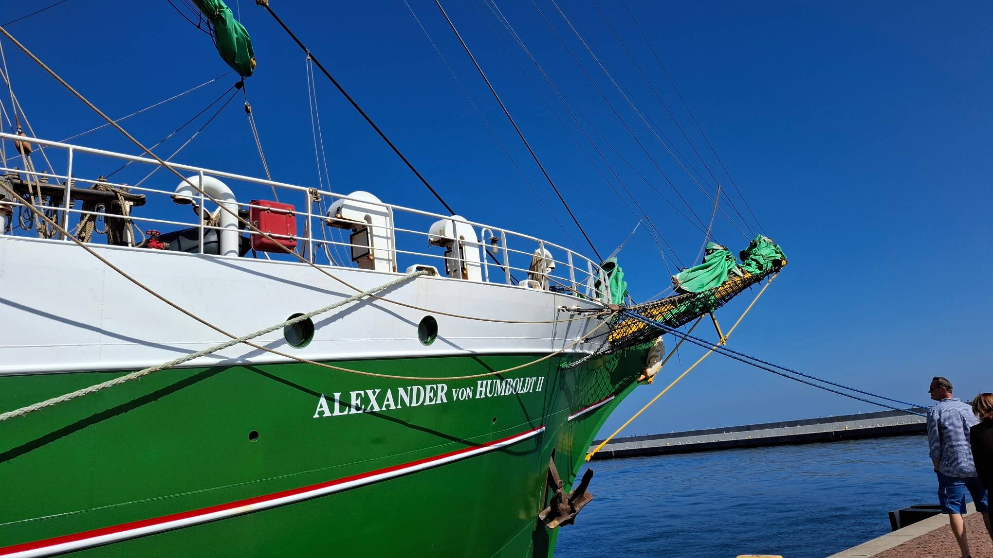 Dziobowa część zielono-białego kadłuba żaglowca Alexander von Humboldt II z wyraźnym białe napisem z nazwą jednostki. W tle widoczny jest port w Gdyni, błękitne niebo oraz przechodzący blisko brzegu ludzie