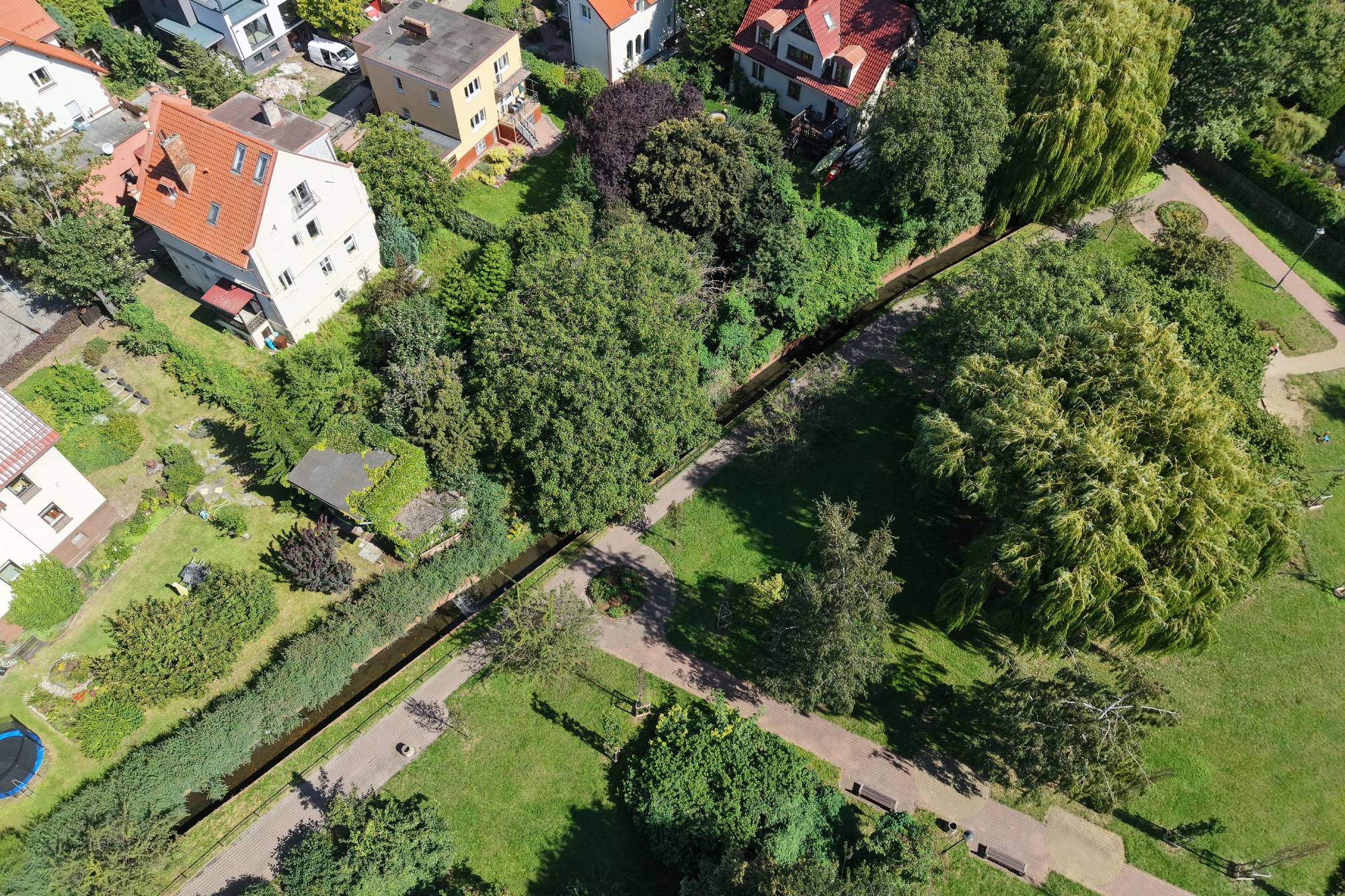 Zdjęcie lotnicze wykonane w słoneczny dzień przedstawia fragment parku sąsiadującego z zabudową mieszkalną. Przez środek kadru, diagonalnie, przebiega koryto Potoku Strzyża, w większości ukryte pod gęstą koroną drzew. Wzdłuż potoku wiją się alejki spacerowe, a teren jest zagospodarowany trawnikami i licznymi nasadzeniami. Po lewej stronie widoczne są dachy domów jednorodzinnych i wielorodzinnych