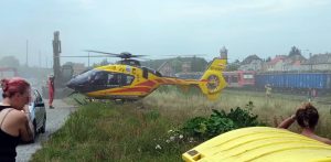 Żółto-czerwony śmigłowiec Lotniczego Pogotowia Ratunkowego z widocznym numerem rejestracyjnym SP-HXW stojący na trawiastym poboczu tuż przy torach kolejowych. W tle widoczny jest czerwony pociąg pasażerski, wagony z drewnem oraz zabudowania miejskie z wieżą. Obok helikoptera stoją świadkowie oraz ratownik medyczny w czerwonym kombinezonie. Wypadek na torach w Miastku. Pociąg potrącił mężczyznę