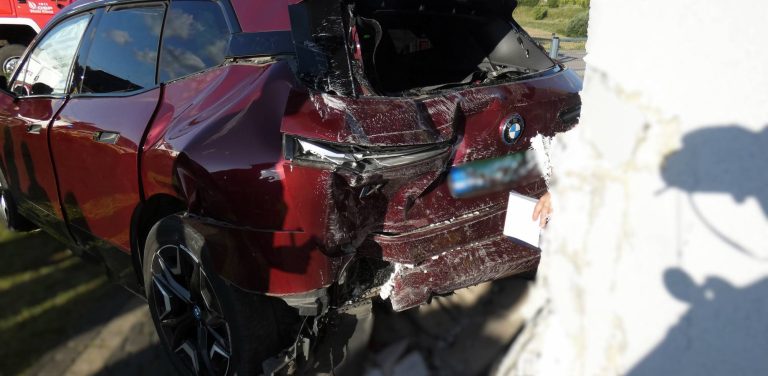 Mocno rozbity tył bordowego samochodu marki BMW po uderzeniu w białą ścianę budynku. Widoczne są silne wgniecenia karoserii, stłuczona tylna szyba oraz uszkodzony zderzak
