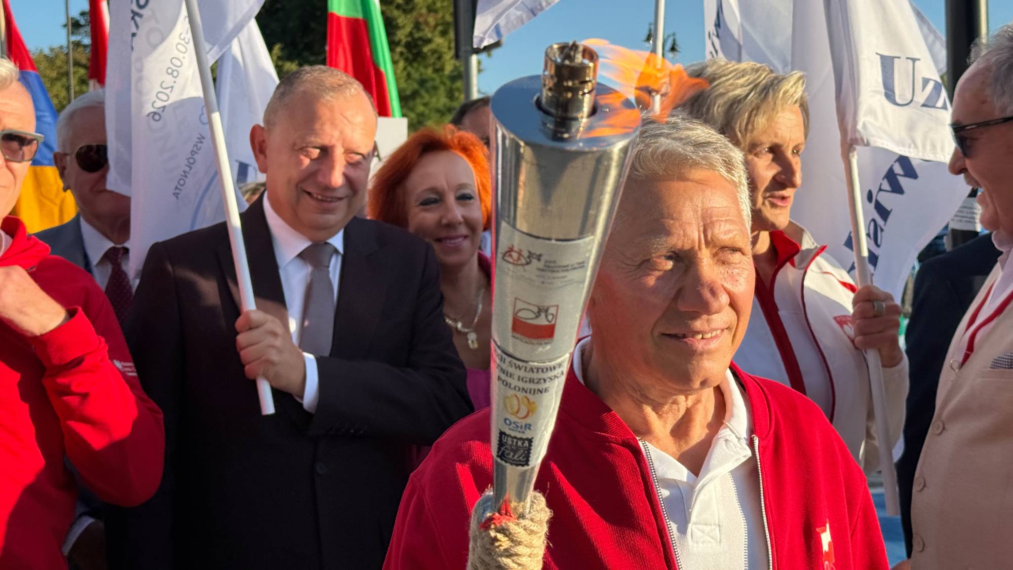 Uroczyste zapalenie i przekazanie ognia olimpijskiego podczas ceremonii otwarcia XXII Światowych Letnich Igrzysk Polonijnych w Ustce. Na pierwszym planie widoczny jest utytułowany olimpijczyk Kazimierz Adach (brązowy medalista w boksie z Moskwy z 1980 roku), który z uśmiechem trzyma płonącą pochodnię igrzysk. W tle towarzyszą mu inni sportowcy, w tym lekkoatletka Barbara Madejczyk oraz zaproszeni goście i uczestnicy wydarzenia