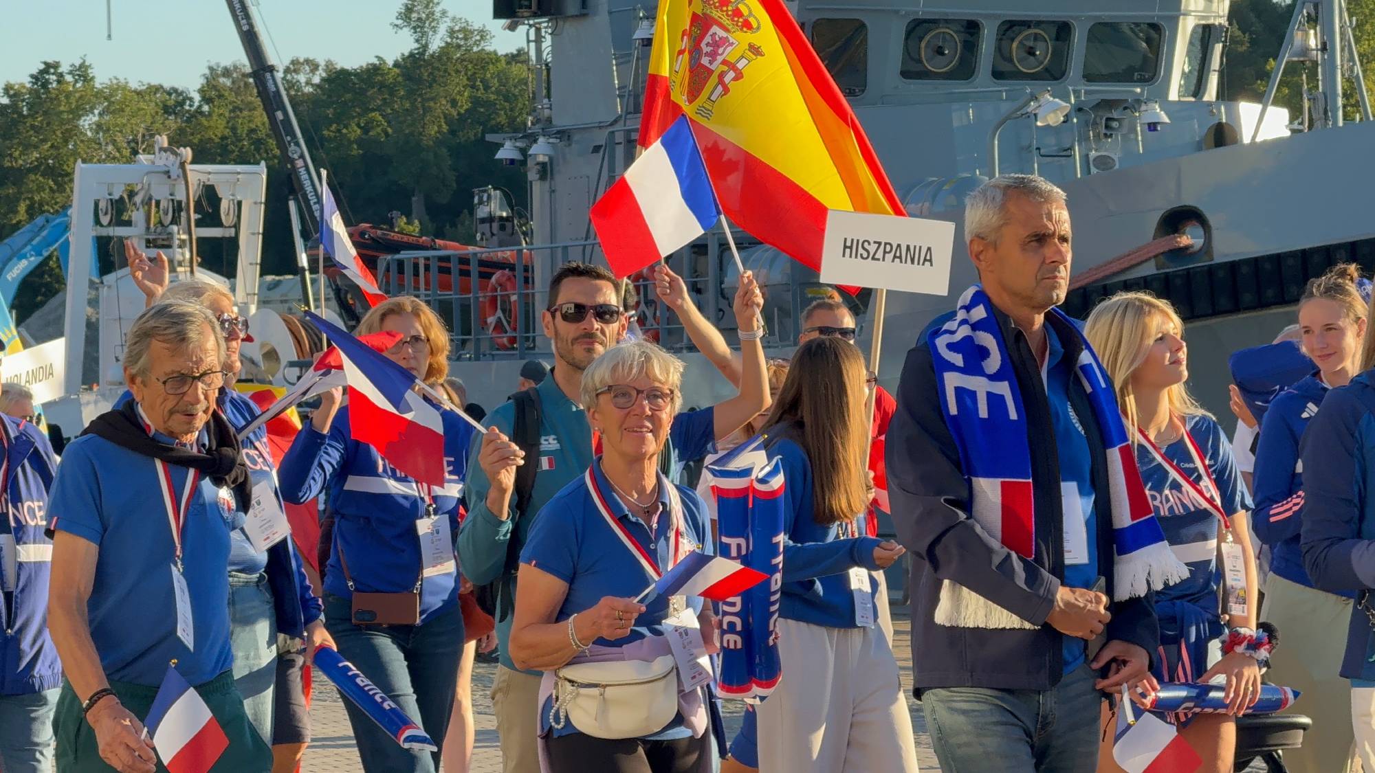Grupa uczestników XXII Światowych Letnich Igrzysk Polonijnych zgromadzona w porcie w Ustce. Na pierwszym planie widoczni są dorośli w niebieskich koszulkach z francuskimi akcentami i szalikami, trzymający małe flaga Francji. W tle ktoś unosi dużą flagę Hiszpanii oraz tablicę z napisem "HISZPANIA", a za nimi góruje szary kadłub statku marynarki wojennej