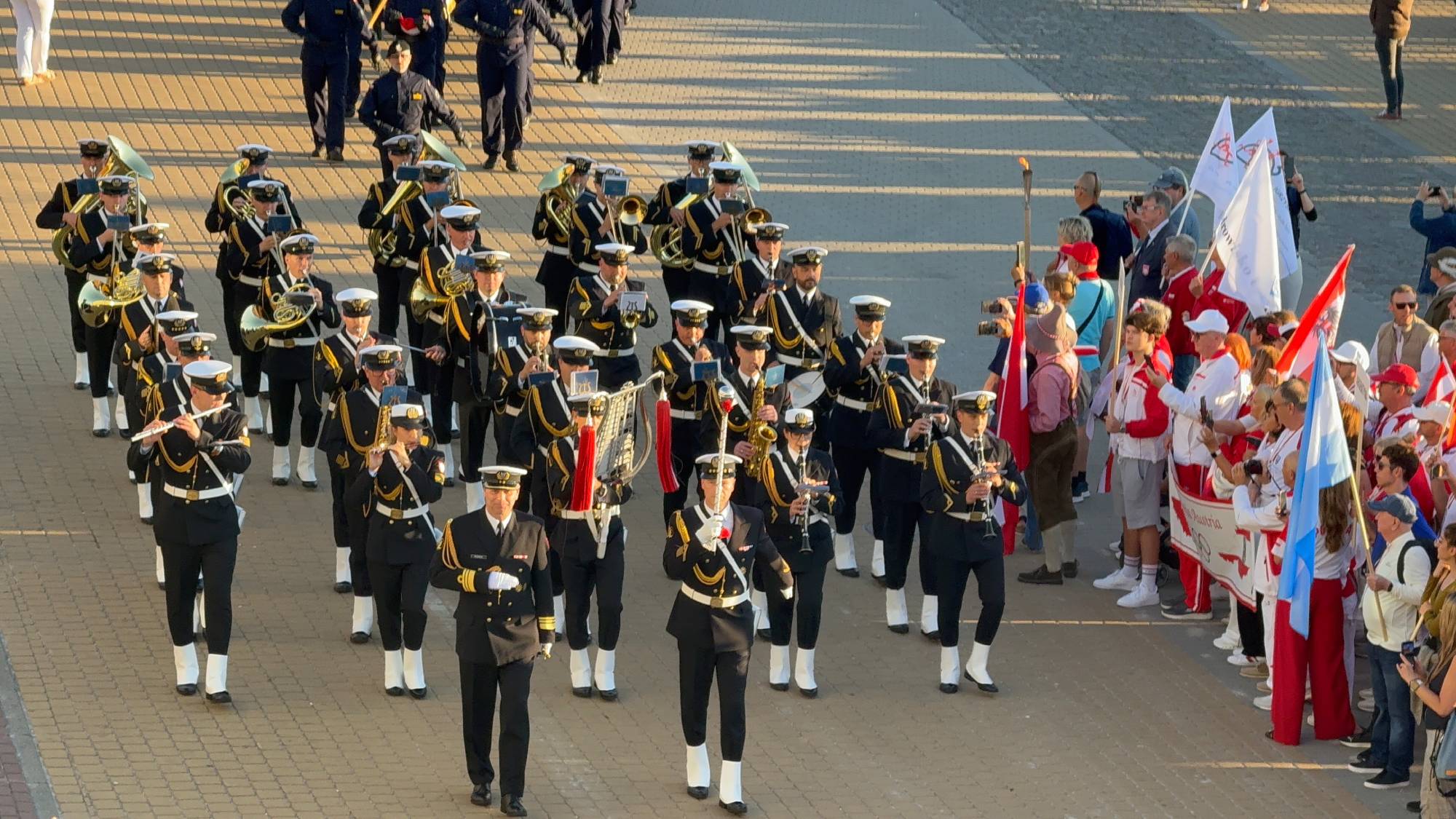 Orkiestra Reprezentacyjna Marynarki Wojennej ubrana w galowe granatowe mundury z białymi czapkami i elementami zdobniczymi maszeruje w szyku po wybrukowanym placu. Muzycy grają na instrumentach dętych. Po prawej stronie stoi szpaler obserwatorów i uczestników igrzysk trzymających flagi oraz znicz olimpijski