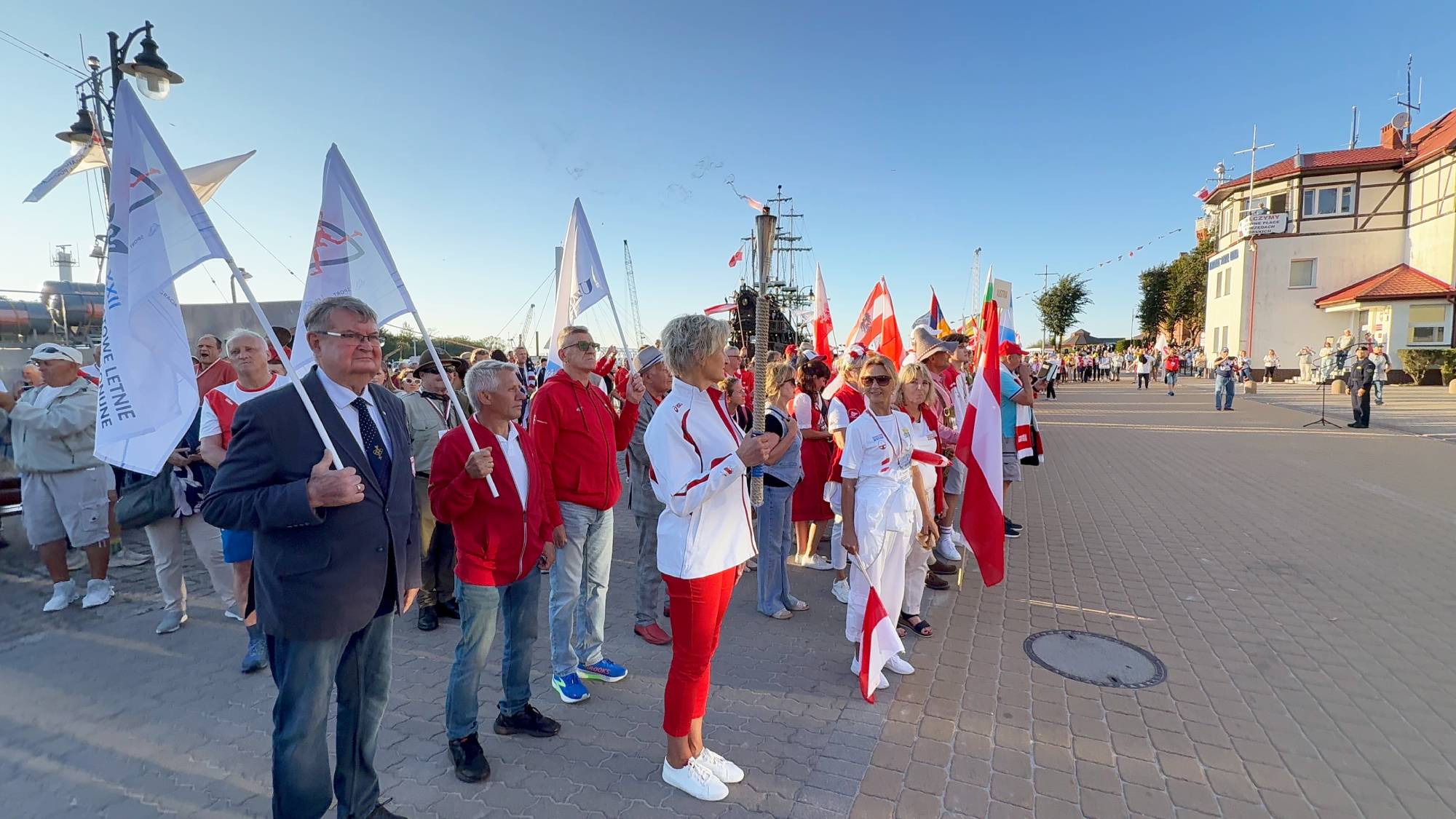Grupa sportowców i olimpijczyków zgromadzona na nabrzeżu w Ustce podczas ceremonii otwarcia igrzysk polonijnych. Na środku stoi kobieta w białej kurtce i czerwonych spodniach (lekkoatletka Barbara Madejczyk) trzymająca zapaloną pochodnię ze zniczem olimpijskim. Obok niej stoją inni uczestnicy w strojach sportowych i garniturach, trzymający flagi narodowe oraz białe flagi z logo igrzysk