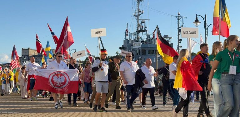 Grupa sportowców i uczestników XXII Światowych Letnich Igrzysk Polonijnych idzie w uroczystej paradzie przez port w Ustce. Ludzie niosą polskie flagi, narodowe barwy oraz tablice z nazwami państw (m.in. "Niemcy", "Łotwa", "Mołdawia"). Na pierwszym planie widoczny jest mężczyzna trzymający biało-czerwoną flagę z godłem i napisem "SC Polonia Hannover", a w tle znajduje się zacumowany statek marynarki wojennej