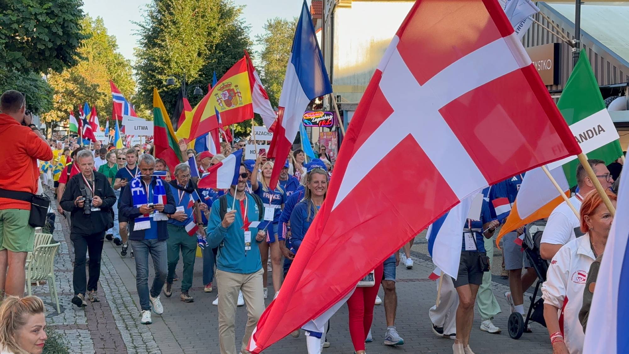 Uczestnicy paradujący ulicą w ramach Polonijnych Igrzysk Olimpijskich. Na pierwszym planie po prawej stronie widoczna jest duża flaga Danii (czerwona z białym krzyżem), a obok niej flaga Francji. W głębi tłum ludzi niesie flagi różnych państw oraz tablice z nazwami krajów, takimi jak "Litwa" i "Finlandia"