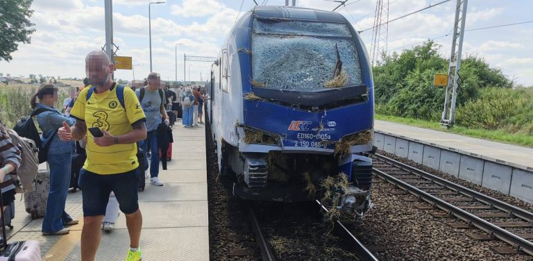 Przód niebiesko-białego pociągu PKP InterCity z mocno popękaną przednią szybą, wgniecionym zderzakiem i wbitymi resztkami słomy. Po lewej stronie, na peronie, stoją i przechodzą pasażerowie z walizkami. Zderzenie pociągu z ciągnikiem