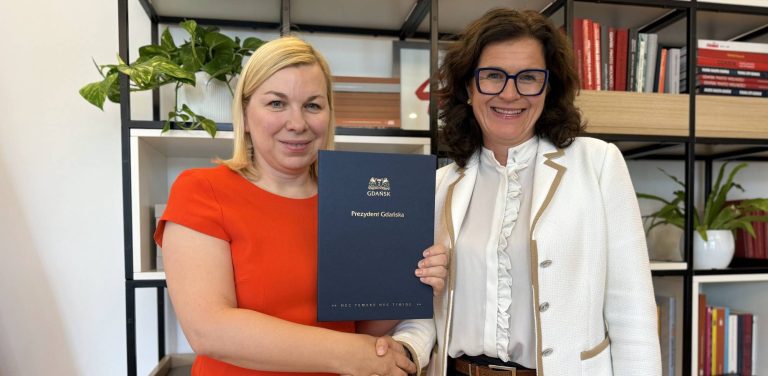 Prezydent Gdańska Aleksandra Dulkiewicz (po prawej, w jasnym żakiecie i okularach) wręcza dokument powołania Celimie Zboromirskiej-Bieńczak (po lewej, w pomarańczowej bluzce), która została nową dyrektor Miejskiego Teatru Miniatura. Obie trzymają wspólnie niebieską teczkę z godłem Gdańska i napisem "Prezydent Gdańska"