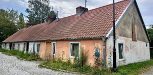 Parterowa chata rybacka (checz rybacka) o spadzistym dachu z czerwoną dachówką i łososiowo-różową oraz białą elewacją przed zbliżającym się remontem. Przed budynkiem, przy brukowanej ulicy, stoi czarna, retro latarnia uliczna. Na ścianie przy oknie widnieje tabliczka adresowa z numerem 17. U podstawy budynku rosną wysokie chwasty