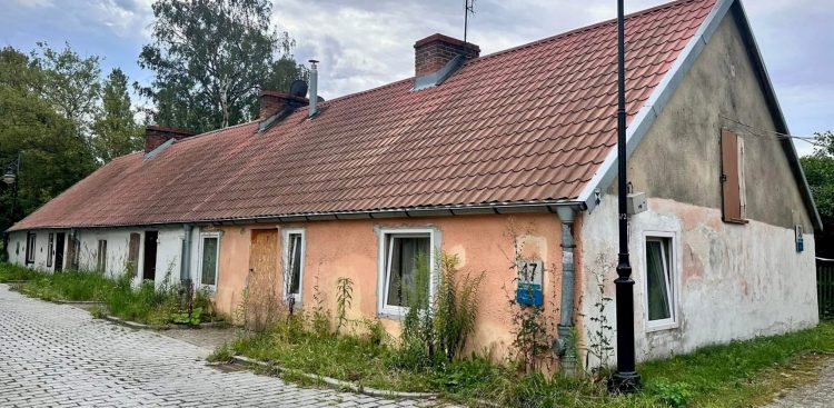 Parterowa chata rybacka (checz rybacka) o spadzistym dachu z czerwoną dachówką i łososiowo-różową oraz białą elewacją przed zbliżającym się remontem. Przed budynkiem, przy brukowanej ulicy, stoi czarna, retro latarnia uliczna. Na ścianie przy oknie widnieje tabliczka adresowa z numerem 17. U podstawy budynku rosną wysokie chwasty