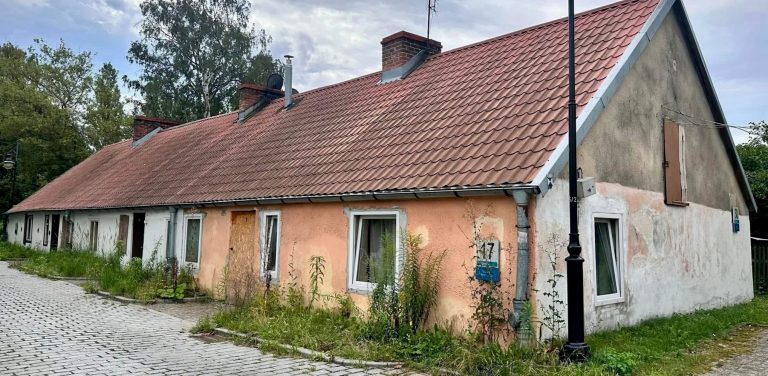 Parterowa chata rybacka (checz rybacka) o spadzistym dachu z czerwoną dachówką i łososiowo-różową oraz białą elewacją przed zbliżającym się remontem. Przed budynkiem, przy brukowanej ulicy, stoi czarna, retro latarnia uliczna. Na ścianie przy oknie widnieje tabliczka adresowa z numerem 17. U podstawy budynku rosną wysokie chwasty