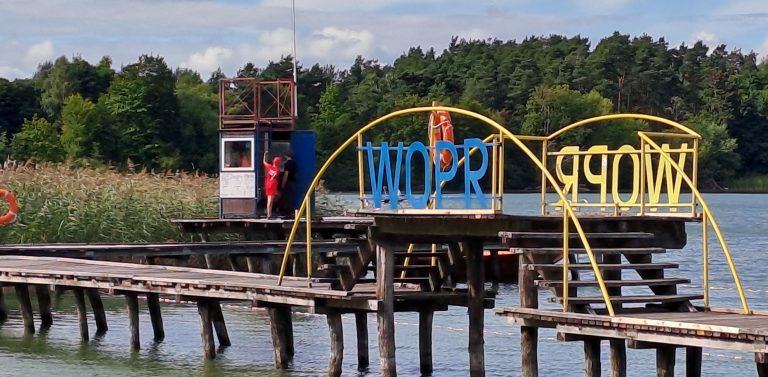 Drewniany pomost nad jeziorem ze stanowiskiem ratowników WOPR i żółto-niebieskimi łukami z napisem „WOPR”