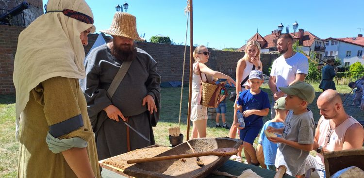 Rekonstruktor w średniowiecznym stroju i słomkowym kapeluszu prezentuje tradycyjne rzemiosło dzieciom i dorosłym podczas pikniku historycznego w Człuchowie