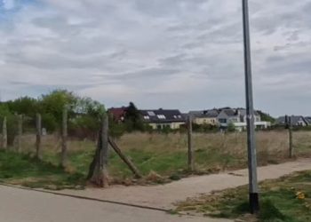 Puste, zielone pole ogrodzone drewnianym płotem w Gdyni Małym Kacku, u zbiegu ulic Strzelców i Żniwnej. W tle widoczne są niskie budynki mieszkalne i zachmurzone niebo. Rada Miasta Gdyni podjęła decyzję o utrzymaniu na tym terenie funkcji rekreacyjno-sportowej