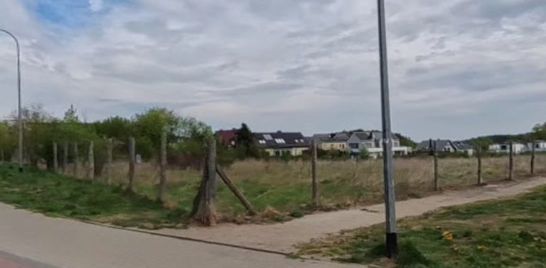 Puste, zielone pole ogrodzone drewnianym płotem w Gdyni Małym Kacku, u zbiegu ulic Strzelców i Żniwnej. W tle widoczne są niskie budynki mieszkalne i zachmurzone niebo. Rada Miasta Gdyni podjęła decyzję o utrzymaniu na tym terenie funkcji rekreacyjno-sportowej