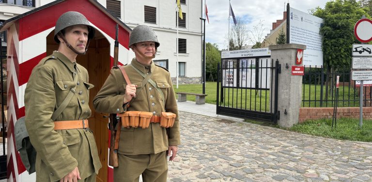 Żołnierze w międzywojennych mundurach stoją przy bramie Muzeum Pomorza Środkowego w Słupsku. Jeden z nich znajduje się tuż obok biało-czerwonej budki wartowniczej, a drugi trzyma karabin