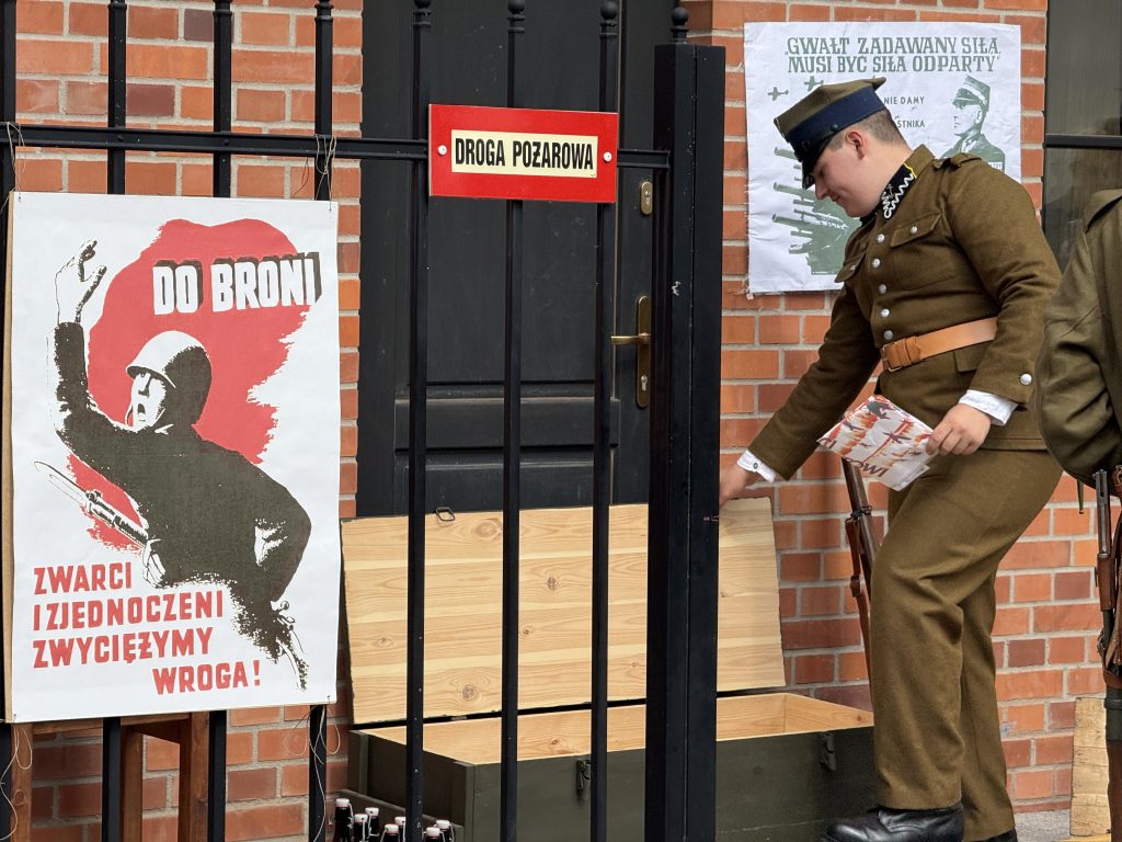 Rekonstruktor w polskim mundurze pochyla się nad drewnianą skrzynią przy czarnej bramie. Obok wiszą historyczne plakaty rekrutacyjne i propagandowe z okresu wojennego