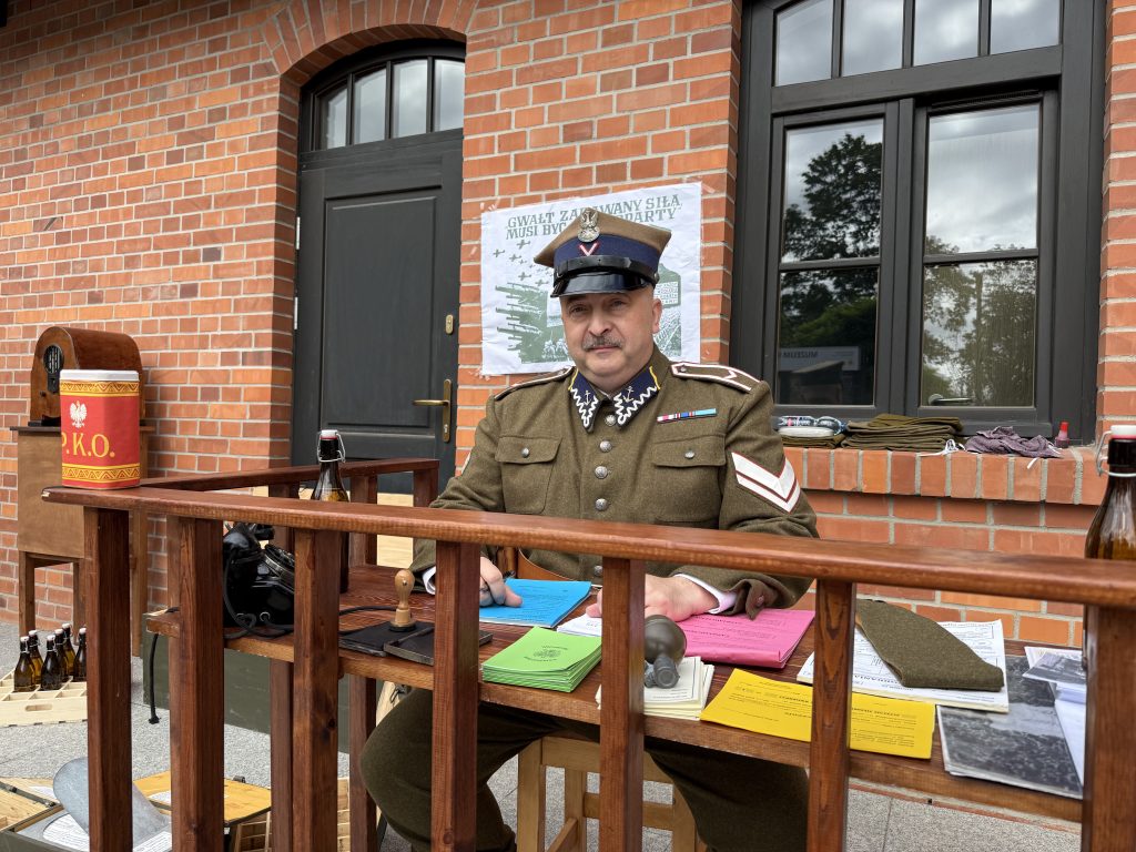 Oficer w mundurze siedzi za drewnianą ladą z dokumentami, pieczątkami i dawnym telefonem. W tle widać ceglaną elewację budynku oraz plakat wojenny