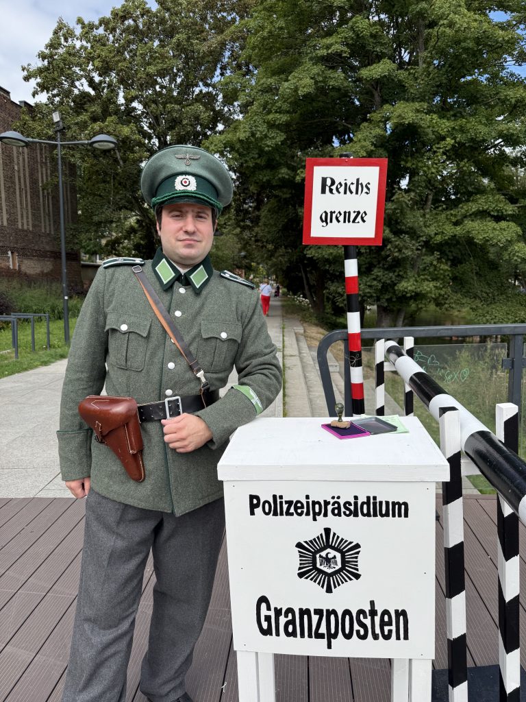 Rekonstruktor w mundurze niemieckiej policji stoi przy punkcie kontrolnym z napisem „Granzposten”. W tle znajduje się biało-czerwony szlaban oraz tablica „Reichsgrenze”