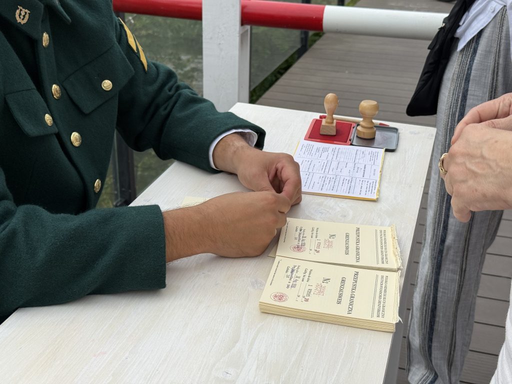 Zbliżenie na stół, przy którym wydawane są historyczne przepustki graniczne „GRENZAUSWEIS”. Na blacie leżą dokumenty, a rekonstruktor stempluje papierowe formularze