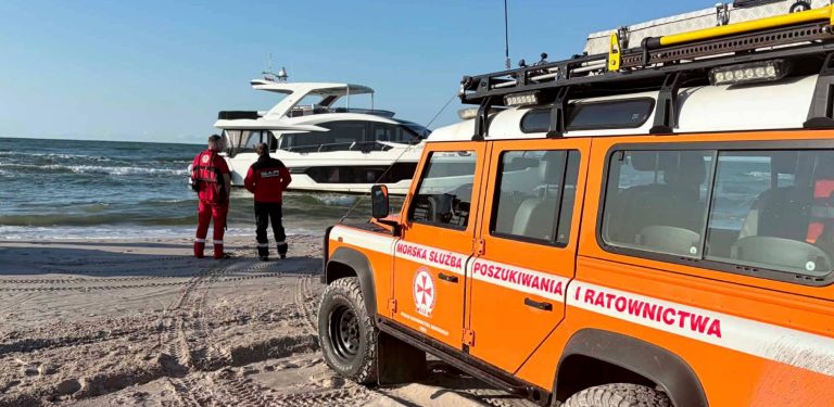 Pomarańczowy samochód terenowy Morskiej Służby Poszukiwania i Ratownictwa (SAR) zaparkowany na piaszczystej plaży. Dwóch ratowników w czerwonych kombinezonach stoi przy brzegu i obserwuje biały jacht motorowy osiadły na płyciźnie