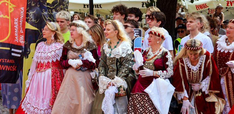 Grupa kobiet w historycznych, bogato zdobionych sukniach renesansowych na tle przechodniów i parasoli ogródków restauracyjnych na Jarmarku św. Dominika