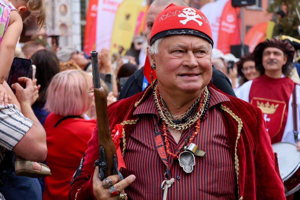 Mężczyzna w przebraniu pirata, w czerwonym welurowym płaszczu i czerwonej chustce z czaszką na głowie, trzymający w ręku starodawną strzelbę na tle tłumu ludzi podczas jarmarku