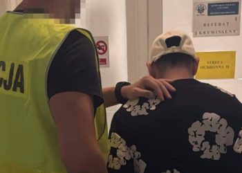 Policjant w odblaskowej kamizelce z napisem "POLICJA" prowadzi zatrzymanego mężczyznę w czarnej wzorzystej koszulce i białej czapce z daszkiem do gabinetu z tabliczką "REFERAT KRYMINALNY"