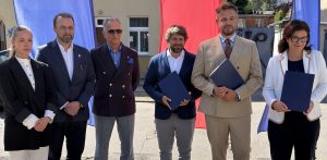 Sześcioro urzędników stoi w rzędzie na zewnątrz budynku na tle niebieskich i czerwonych flag. Trzech mężczyzn i jedna kobieta trzymają w dłoniach granatowe teczki z dokumentami, a pozostałe dwie osoby stoją obok nich. Wszyscy patrzą w obiektyw. Podczas oficjalnej ceremonii władze Gdańska podpisały umowę na budowę drugiego etapu "Kanału Ulgi" dla Potoku Strzyża